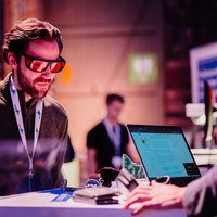 Congresbezoeker met bril kijkt naar demonstratie van onderzoeksproject -- klik op de afbeelding om te vergroten