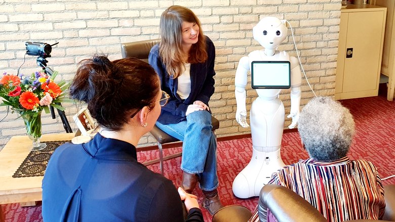 Bewoner van zorginstelling heeft interactie met sociale robot Memo, onder begeleiding van zorgmedewerker en auteur van Wintertuin
