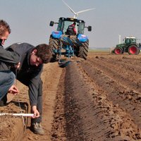 Twee onderzoekers doen bodemonderzoek in een akkerveld met op de achtergrond verschillende tractoren -- klik op de afbeelding om te vergroten