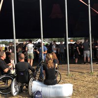 Mensen zitten in een rolstoel of op de rolstoelbank en kijken uit op een mensenmenigte in een festivaltent -- klik op de afbeelding om te vergroten