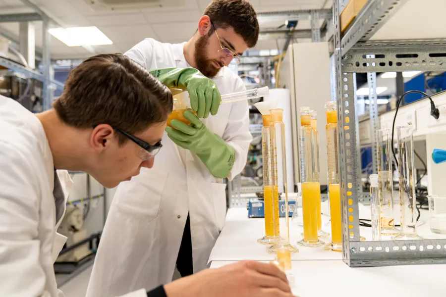 Man giet gele vloeistof in buis, in lab. Andere man kijkt toe. 